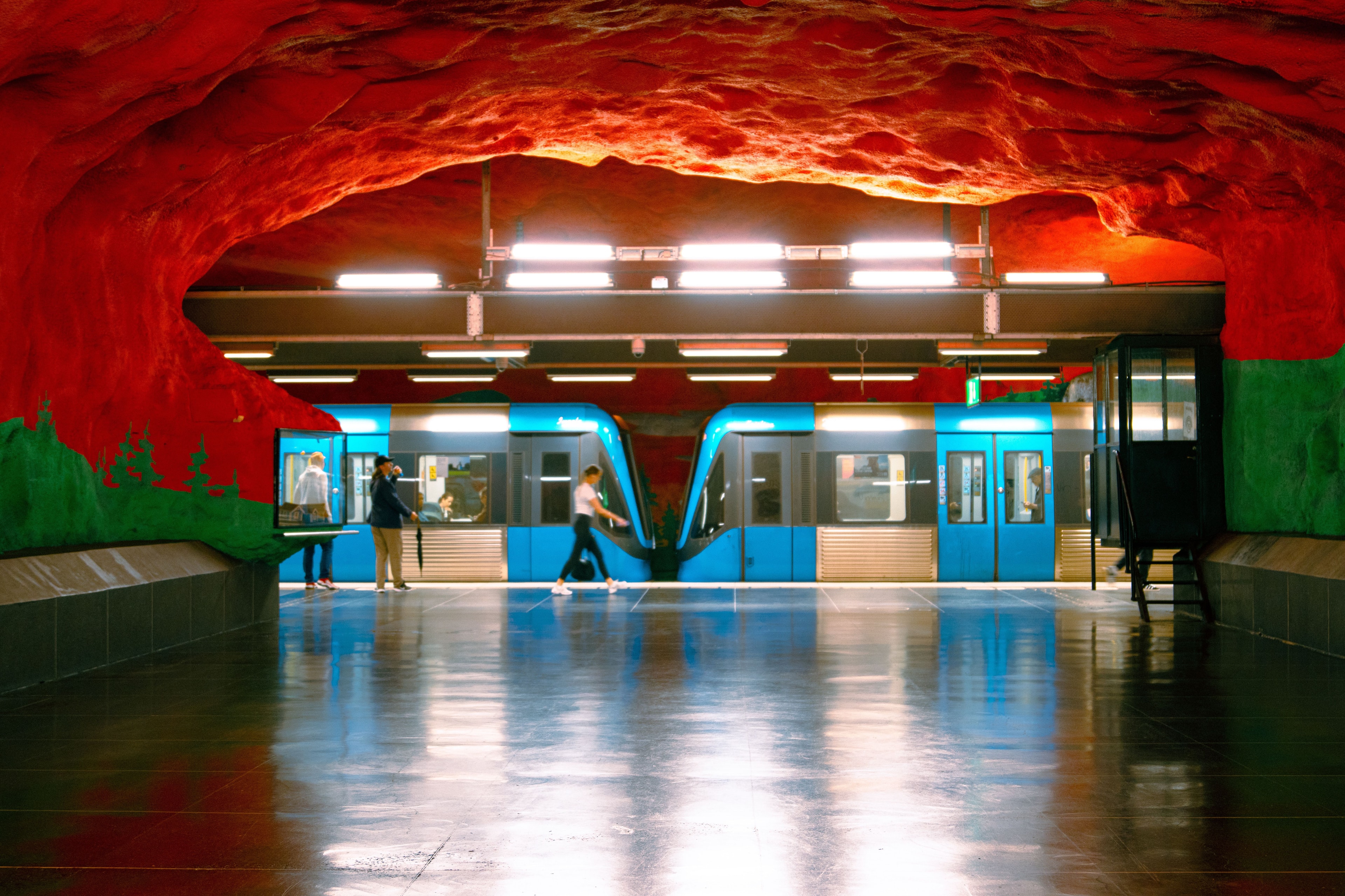 Treno della metro azzurro in una stazione dipinta di rosso e verde, con passeggeri sul marciapiede