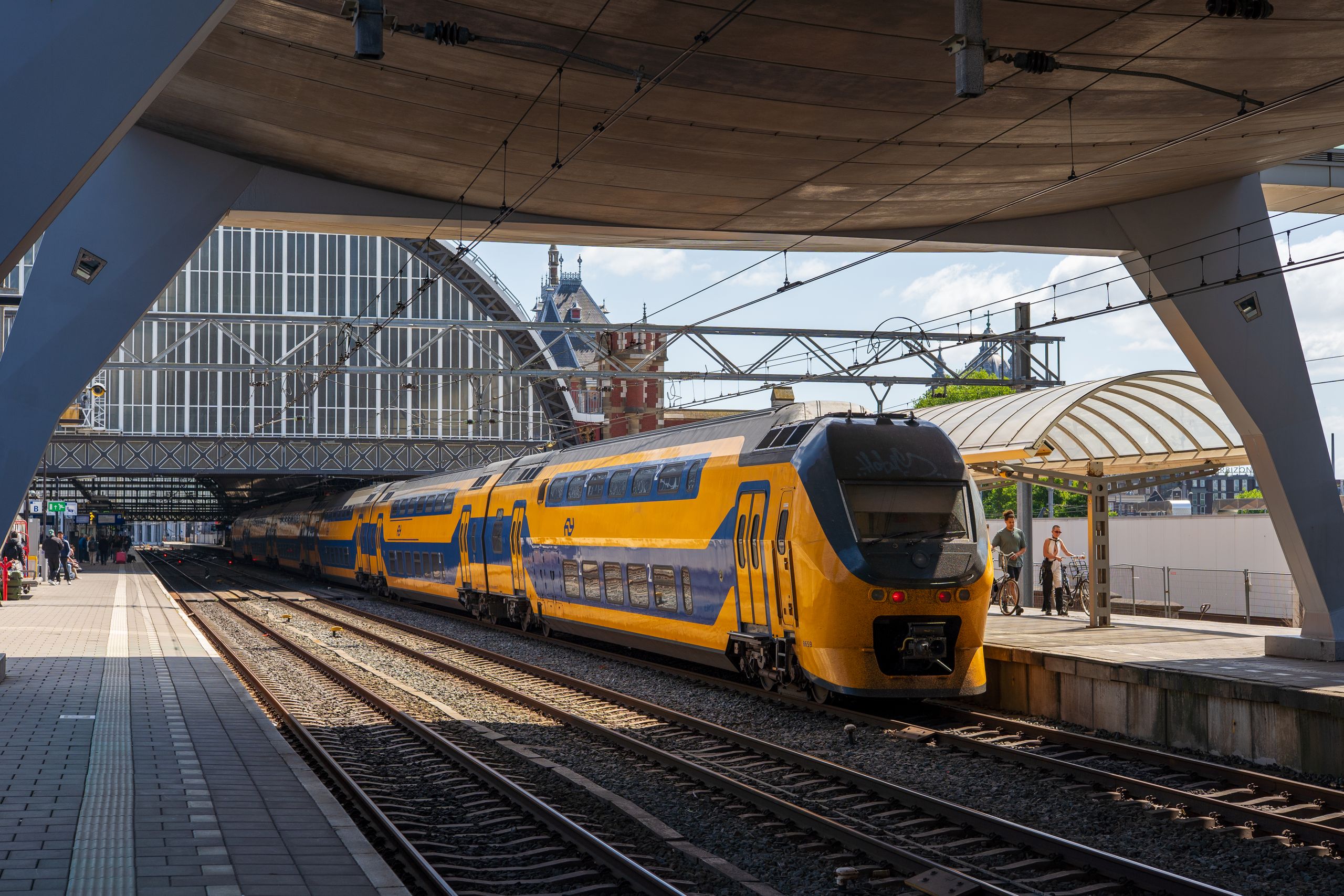 Treno regionale a due piani, giallo e blu, in partenza dalla stazione di Amsterdam Centraal in una giornata di sole