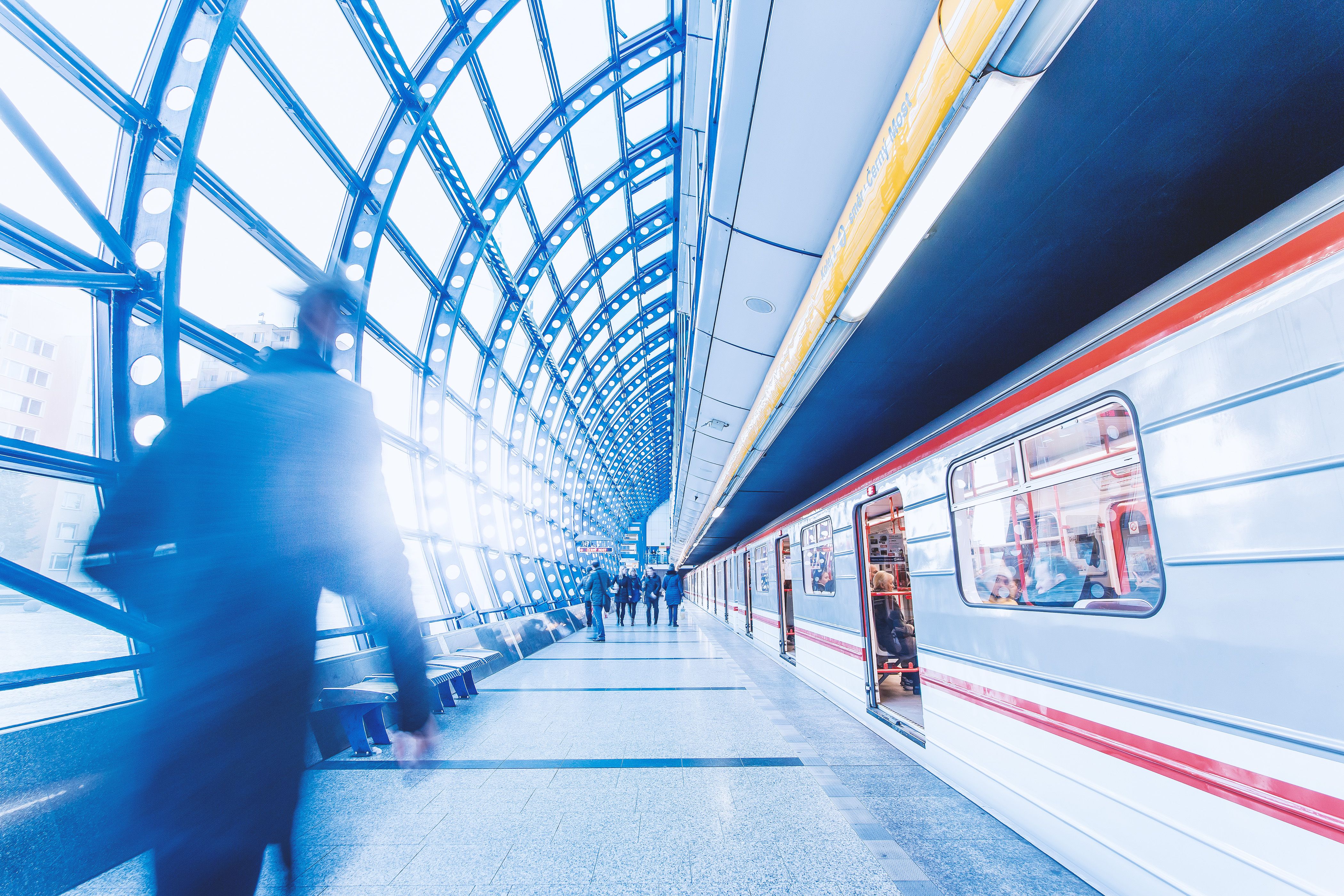 Pendolari in movimento sotto la volta di vetro di una stazione, accanto a un treno fermo in banchina
